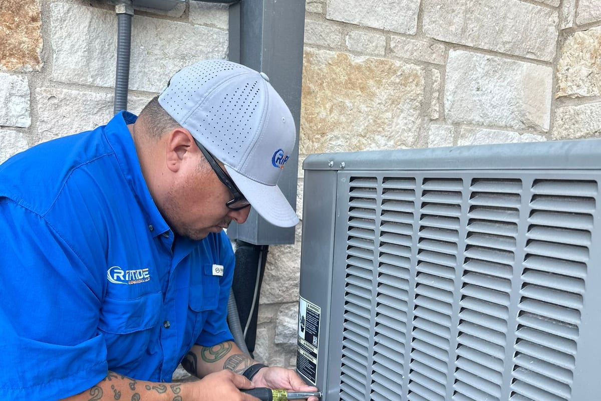 A Riptide Mechanical technician with gauges connected at an outdoor condenser, against the limestone wall of an Austin area home