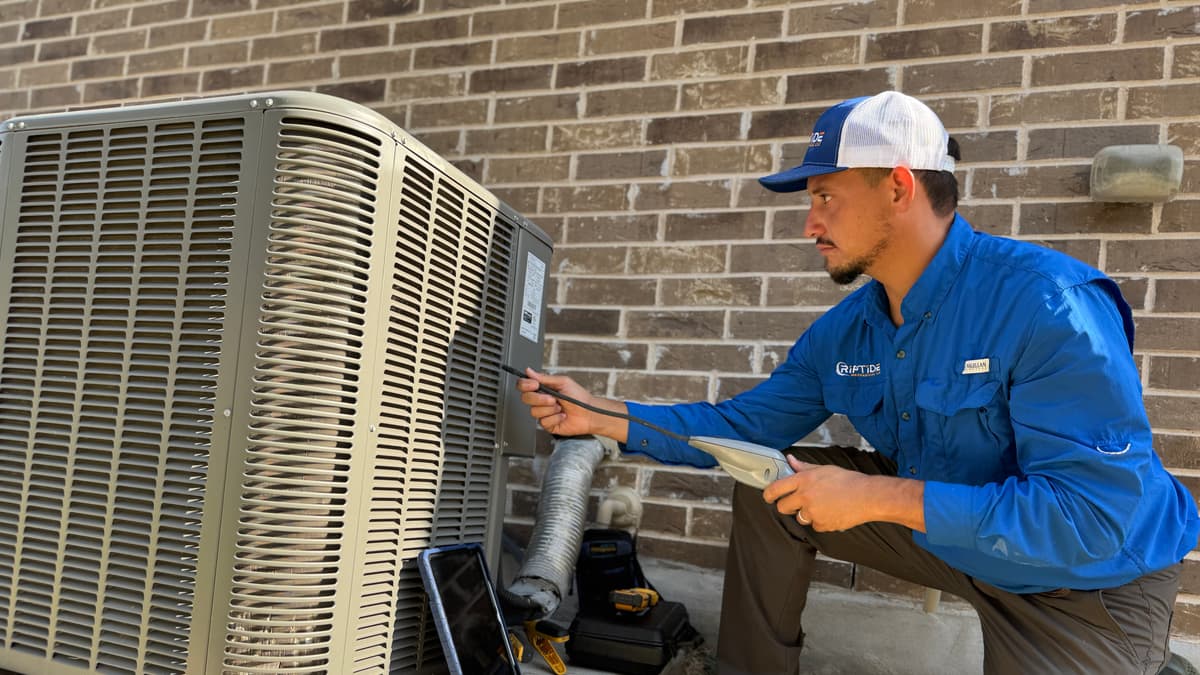 A Riptide Mechanical technician kneeling beside a residential outdoor condenser unit with a probe